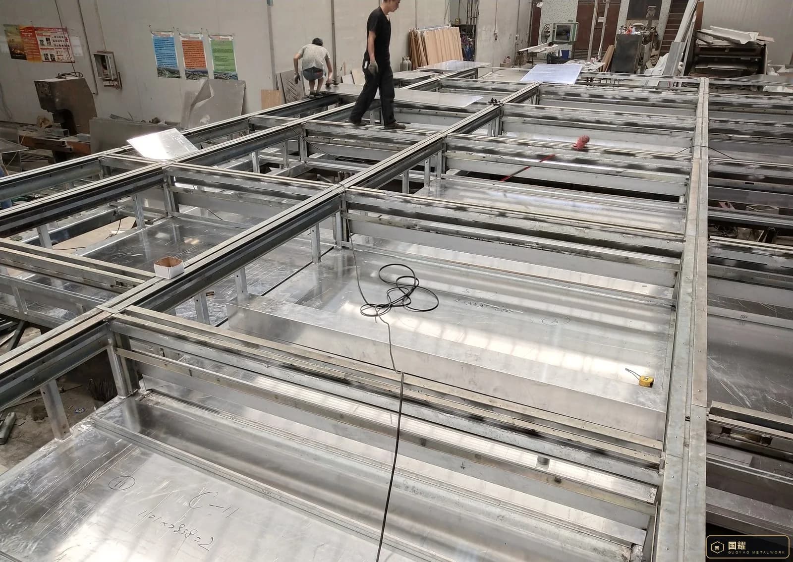 Worker laying a metal skin panel onto a facade sub-frame in a workshop, with several frames in rows behind