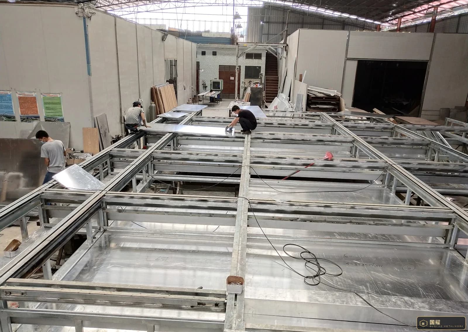 Two workers fitting and checking a flat metal panel on a long facade frame in a workshop