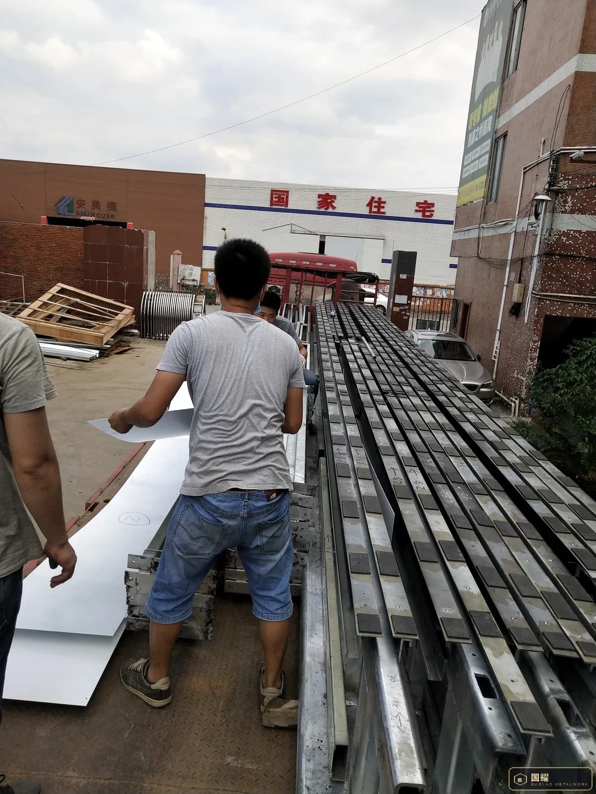Workers loading long facade frames onto a flat rack truck at the factory gate